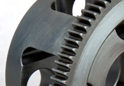 a close-up view of a gear part. It has a dark gray or metallic color with a finely machined surface. The gear teeth are well-defined and exhibit sharp edges, indicative of high precision in manufacturing. There is a central hole, which likely serves as a mounting point to a shaft or similar component. The background is blurred, which brings the gear and its detailed features into focus. This component is probably used in machinery that requires interlocking gears, possibly in automotive, industrial equipment, or robotics applications where precise motion is essential.