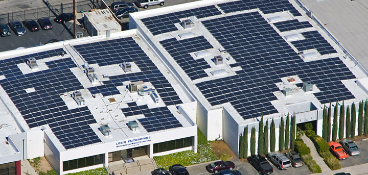 an aerial view of the Lees Enterprise buildings with a significant number of solar panels installed on its roof. The panels are laid out in neat rows, covering most of the available rooftop space, which suggests a commitment to sustainable energy practices. Air conditioning units are visible on the rooftop, interspersed among the solar panels. The building is labeled "LEE'S ENTERPRISE," indicating the name of the business. The parking lot adjacent to the building is partially filled with cars, indicating that the photo was taken during business hours. This setup reflects an investment in renewable energy, likely to reduce energy costs and the environmental footprint of the business.